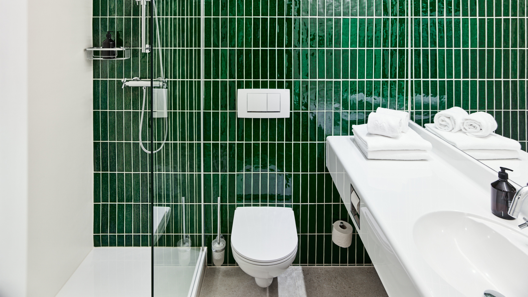 Modernes Badezimmer mit grünen Kacheln, weißer Toilette und Waschtisch. Handtücher und Dusche sind ebenfalls sichtbar.