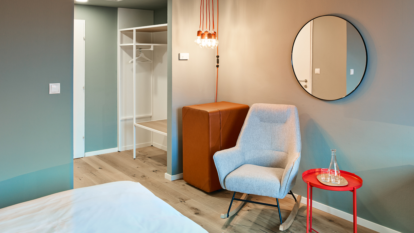 Innenraumfoto zeigt Schaukelstuhl, Beistelltisch und Kleiderschrank. Das runde Spiegelbild des Raumes ist an der Wand sichtbar.