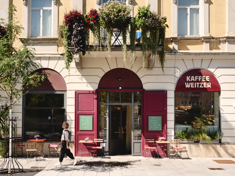 Außenaufnahme des Kaffee Weitzer mit blumengeschmücktem Balkon und einer Person, die vorbeigeht.