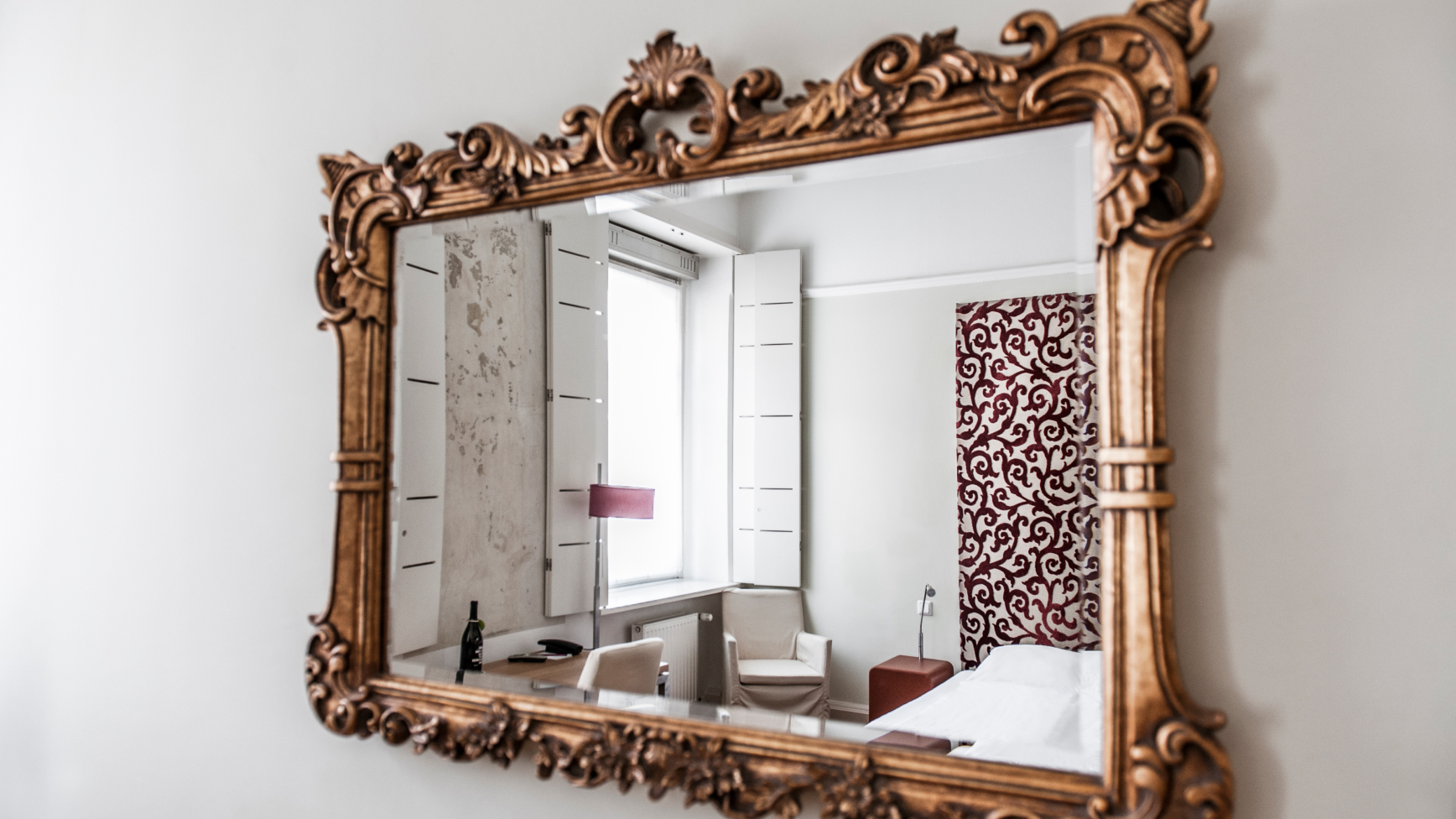 Ein Spiegel mit einem verzierten Goldrahmen, der ein Schlafzimmer mit einem Schreibtisch und einem Bett reflektiert.