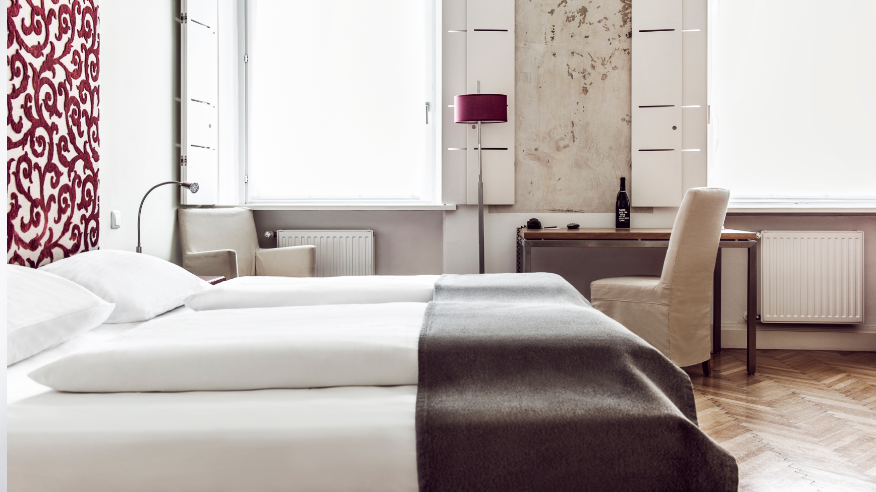 Helles Hotelzimmer mit Doppelbett, Schreibtisch, Stuhl, Lampe und dekorativer Wandgestaltung.