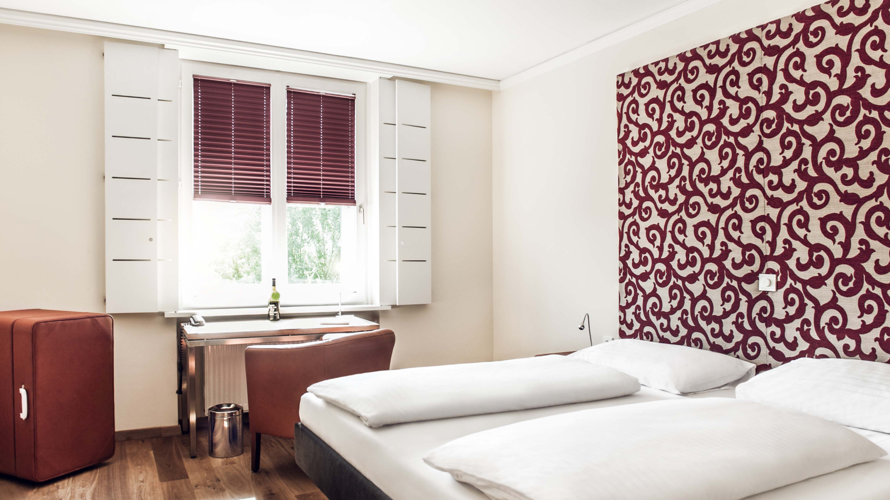 Schlafzimmer mit Doppelbett, Schreibtisch und roter Akzentwand mit Wirbelmuster.