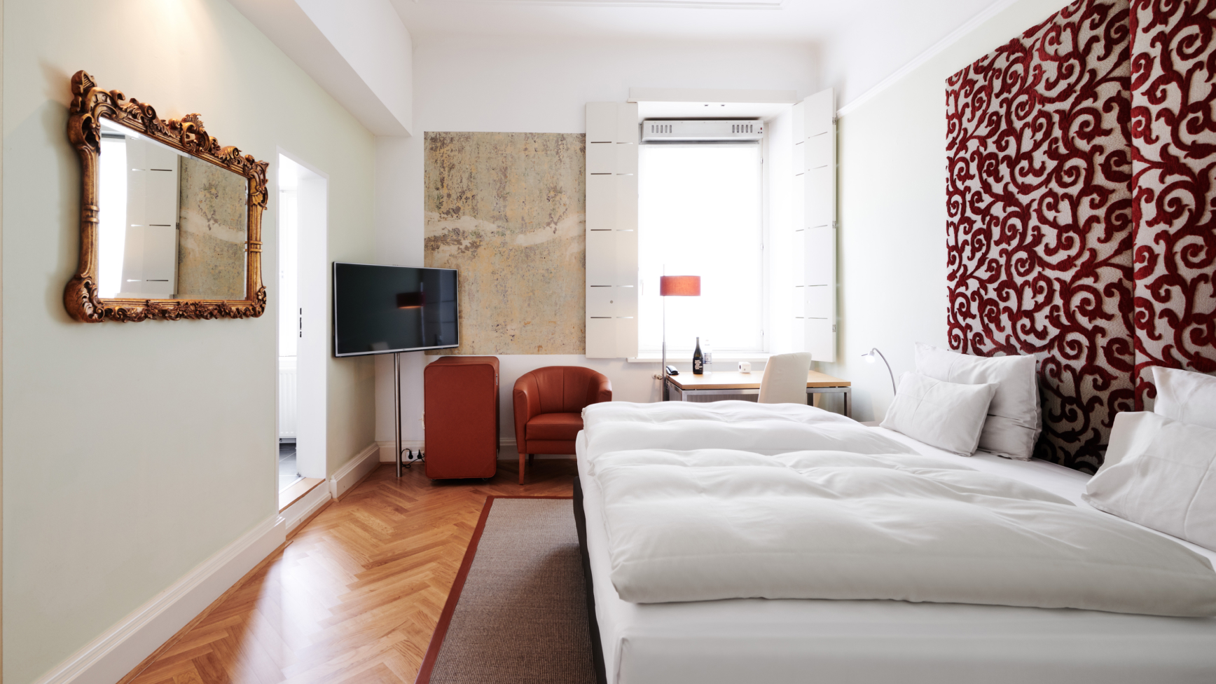 Ein Hotelzimmer mit großem Spiegel, Fernseher, Schreibtisch und Bett mit roter, gemusterter Kopfseite.