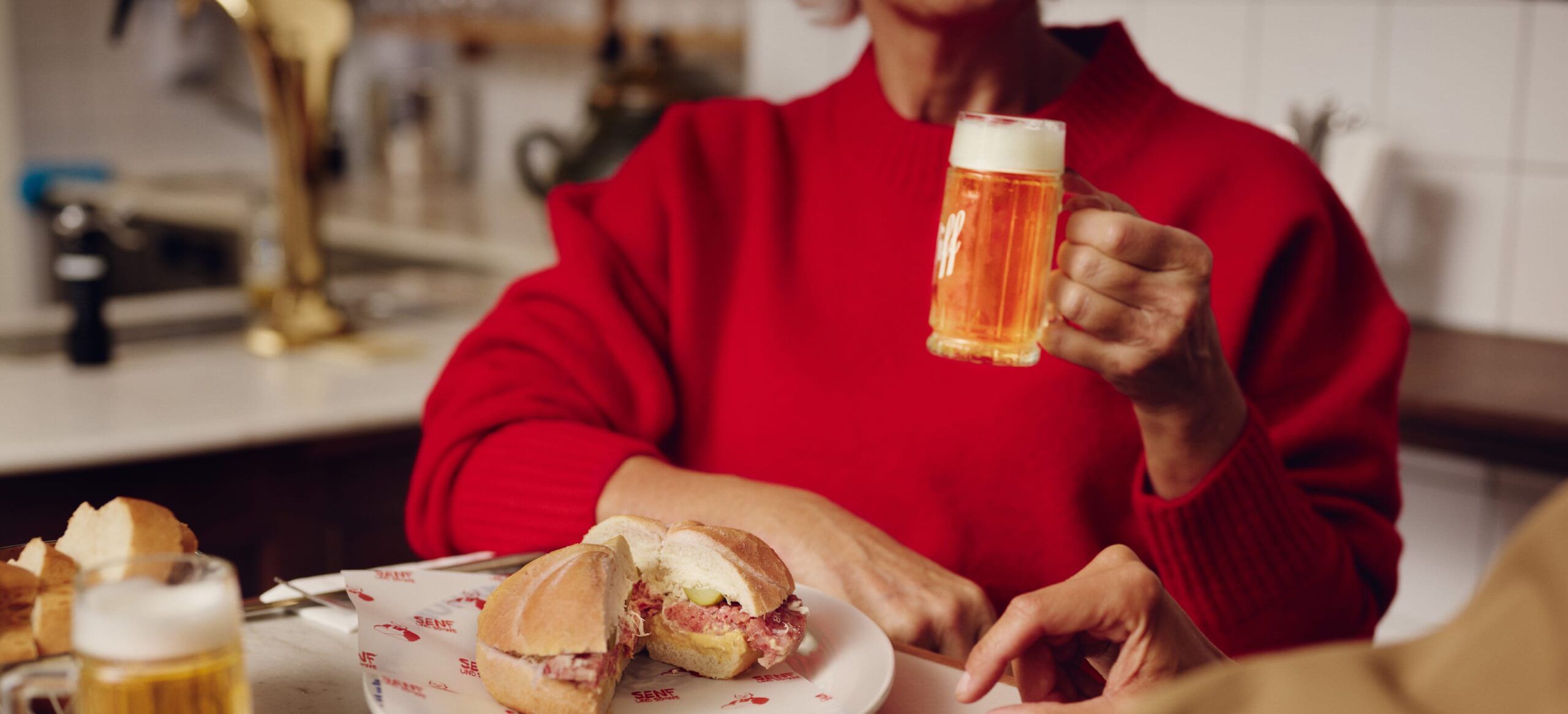 Nahaufnahme einer Person in Rot mit Bier und Sandwich auf einem Tisch.