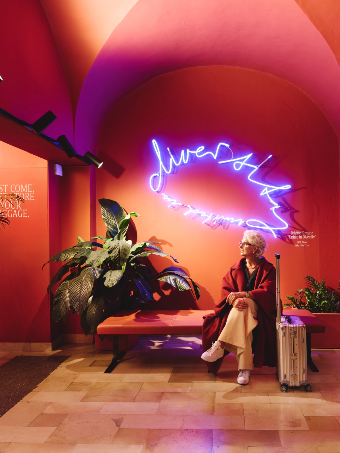 Eine Frau, sitzt mit Koffer auf einer Bank vor einer Neonarbeit von Brigitte Kowanz mit der Aufschrift „United in Diversity“.