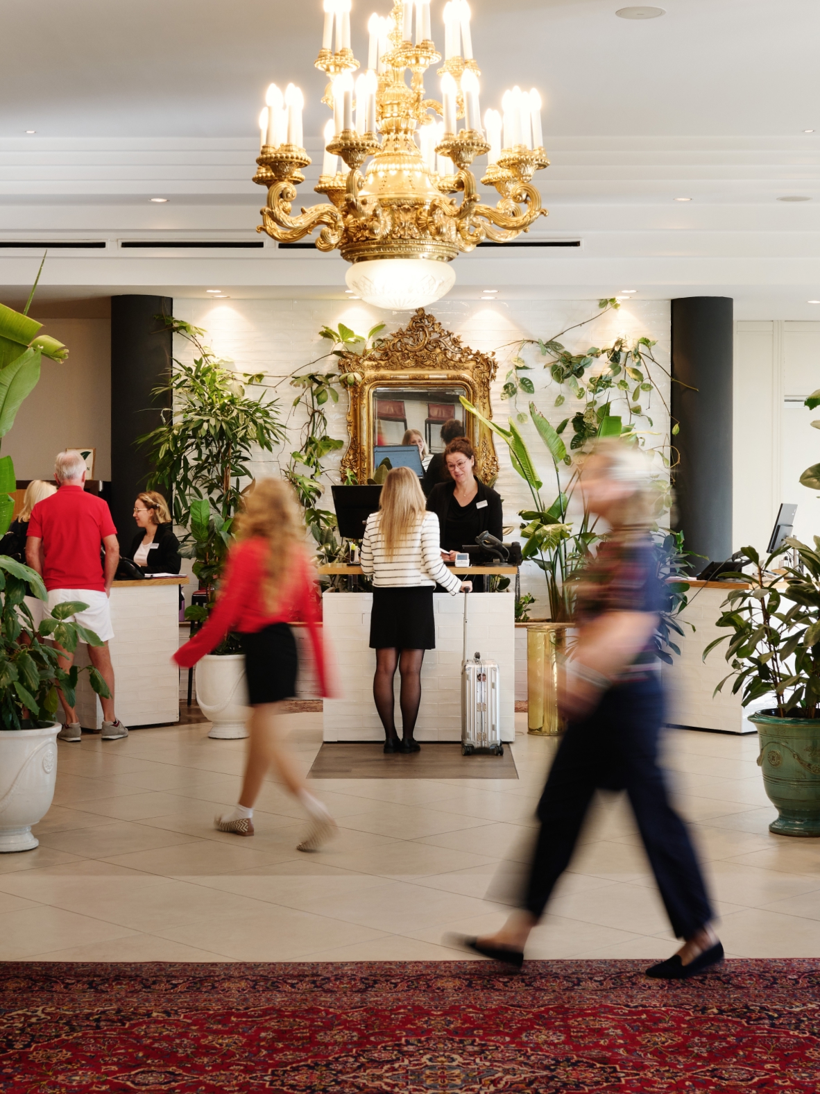 Eine Hotellobby mit einem großen Kronleuchter, einem Spiegel und einer Rezeption mit Gästen.