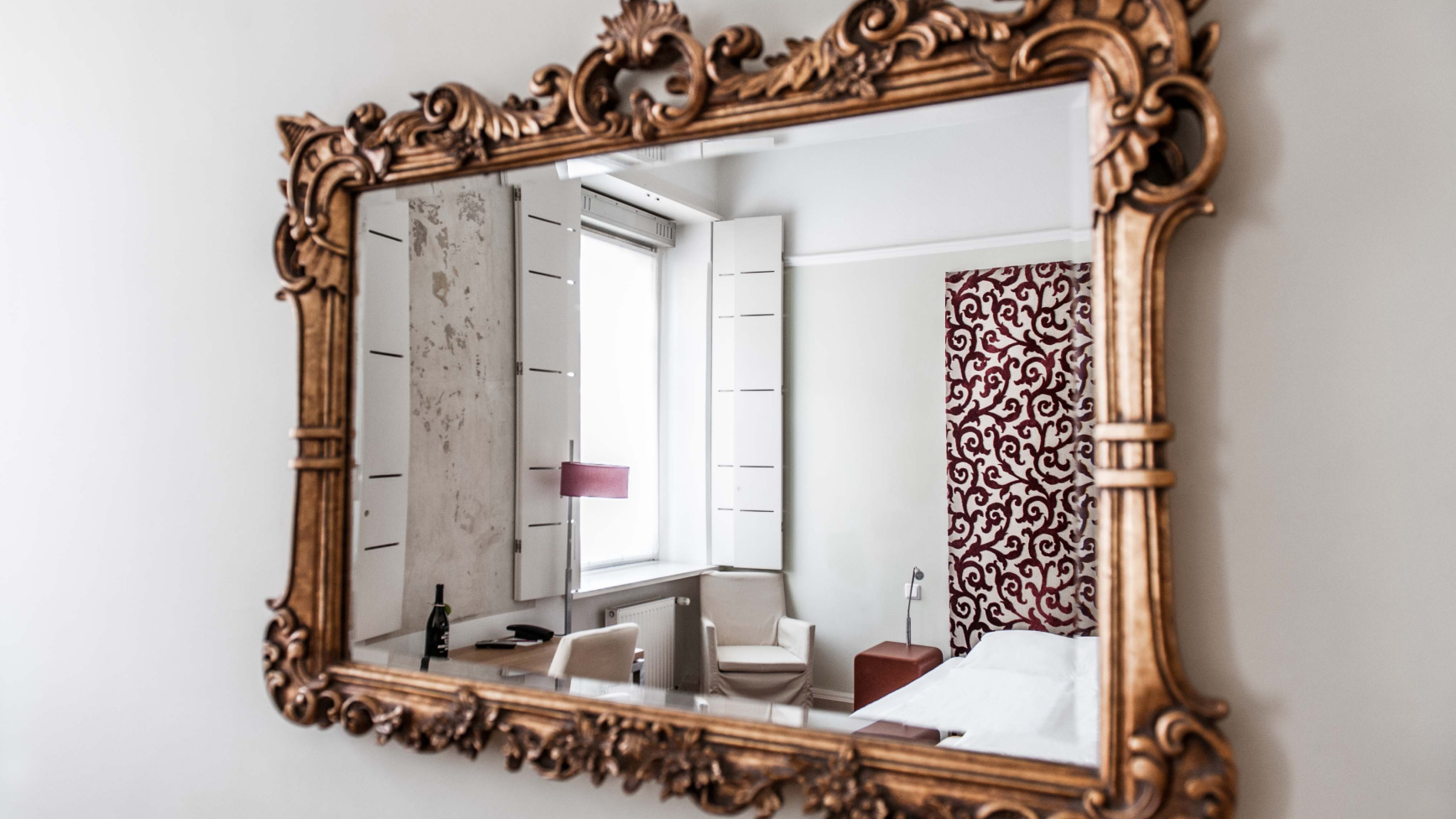 Vergoldeter Spiegel reflektiert ein Schlafzimmer mit Fenster, Schreibtisch und rotem Akzentmuster.