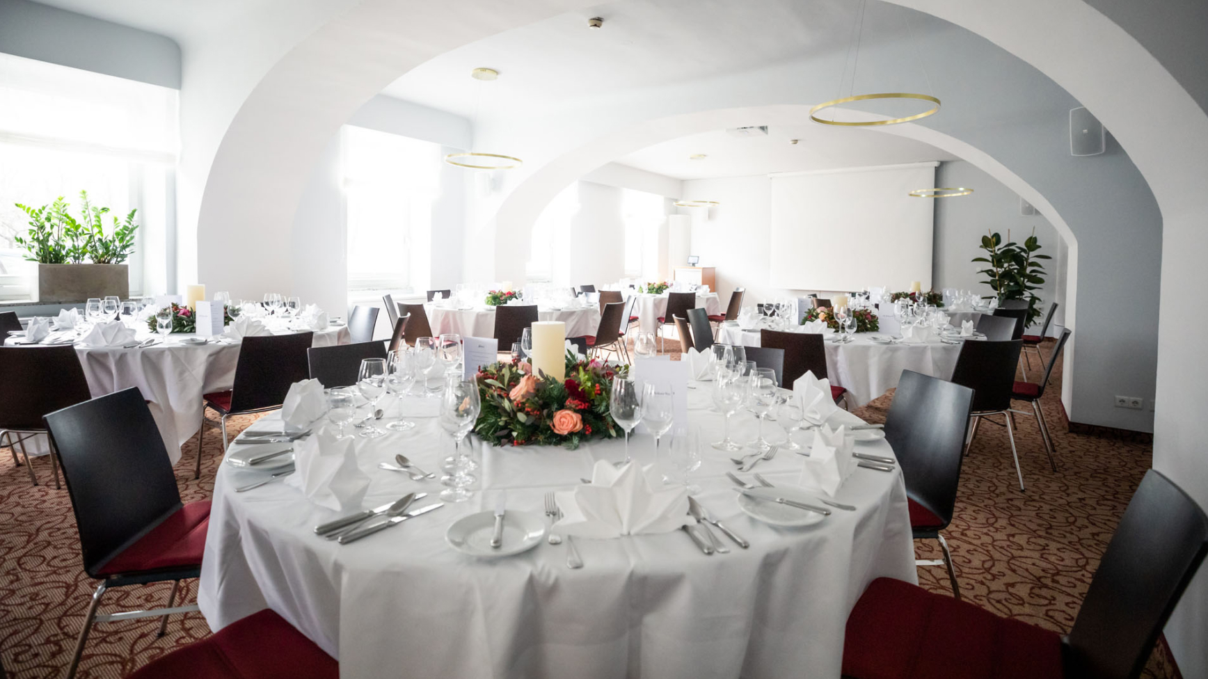 Innenaufnahme eines Veranstaltungsraumes mit runden Tischen, weißer Tischwäsche und Blumengestecken.