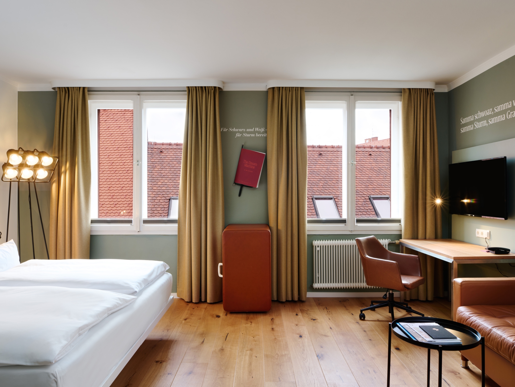 Ein Hotelzimmer mit Bett, Schreibtisch, Sessel und Fenster mit Blick auf rote Ziegeldächer. Zitate an der Wand.