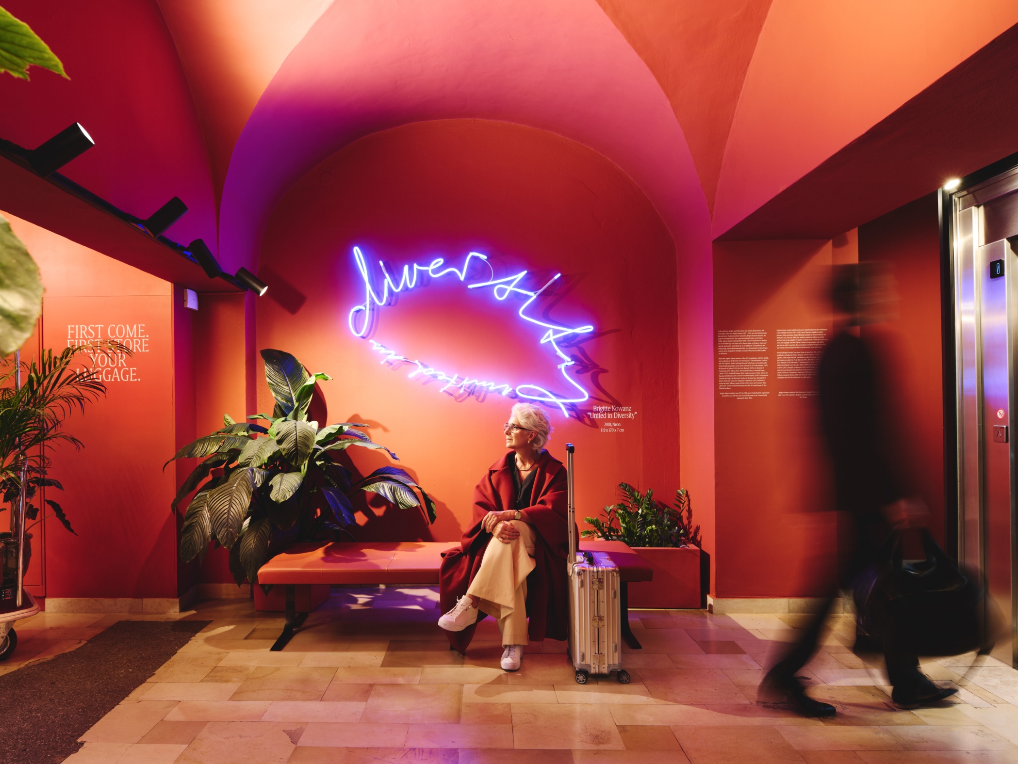 Eine Frau sitzt neben ihrem Koffer unter der Neonbeschriftung „Diversity“ in einem orangefarbenen Hotel.
