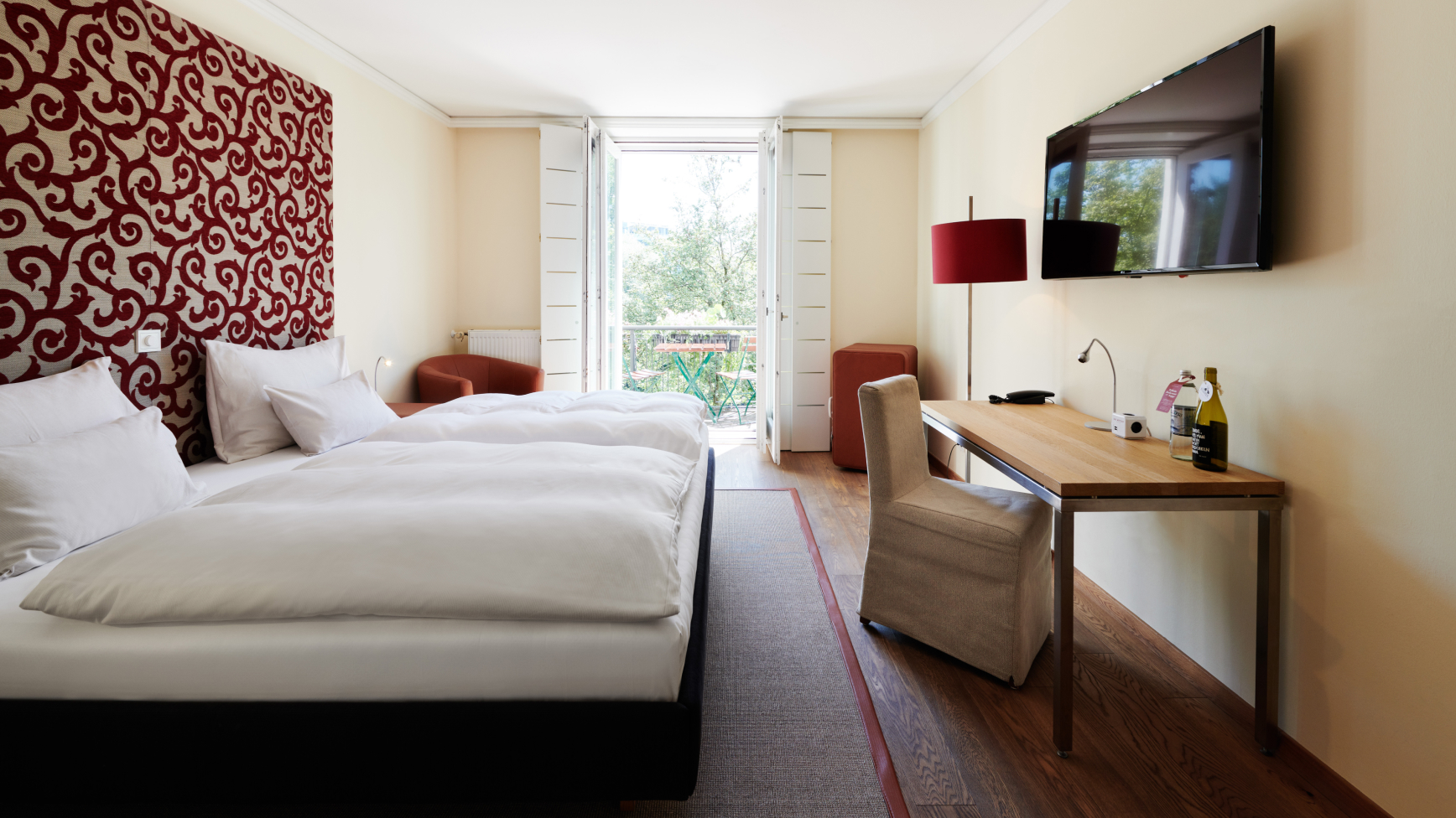 Helles Hotelzimmer mit Doppelbett, Schreibtisch, Fernseher und Balkon mit Blick ins Grüne.