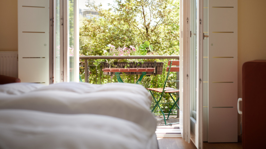 Blick vom Bett auf Balkon mit Tisch und Grün. Offene Türen, entspannende Atmosphäre.