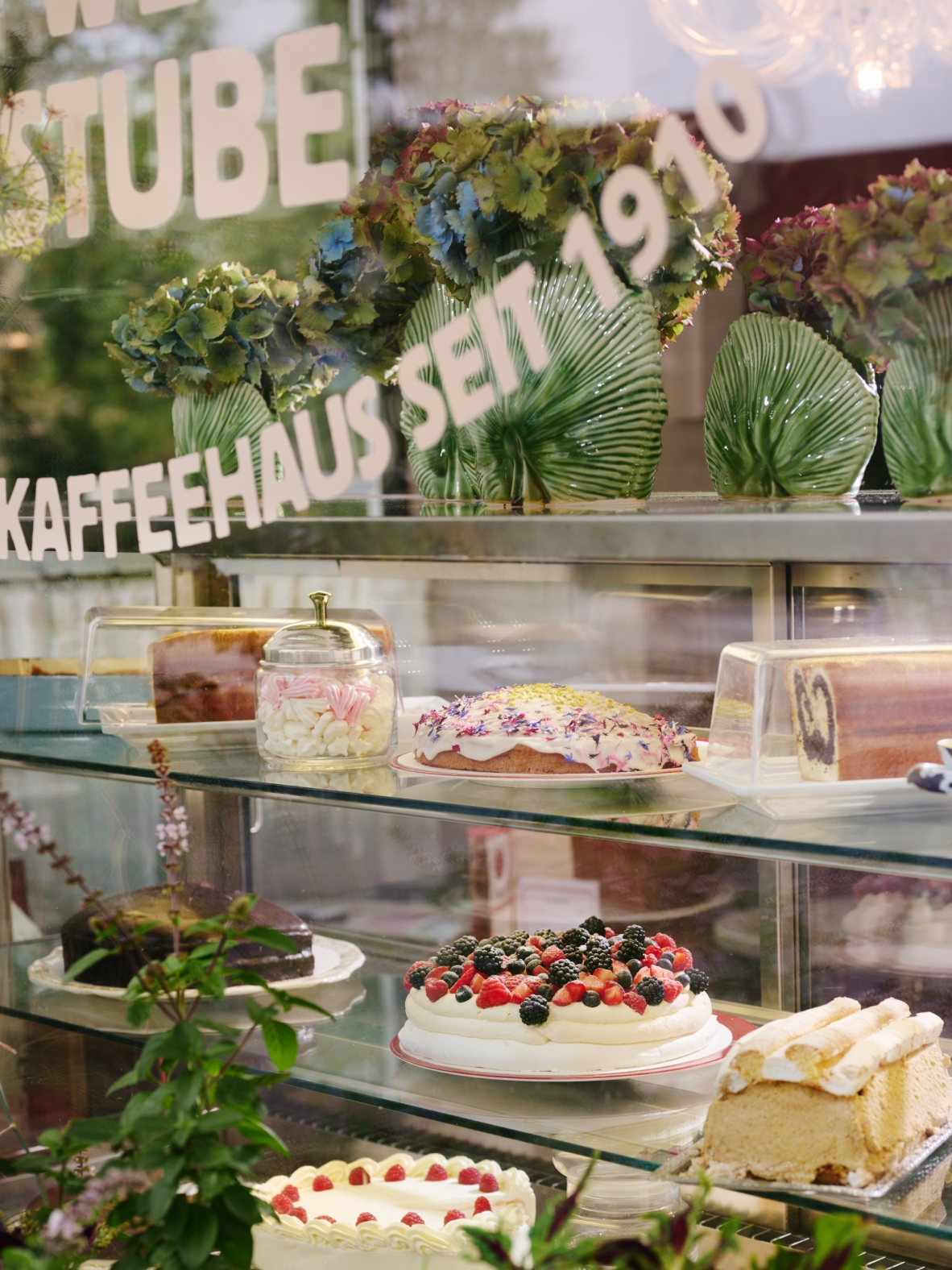 Eine Konditoreiauslage mit Kuchen, Torten und Schriftzug "Kaffeehaus Seit 1910" auf der Scheibe.