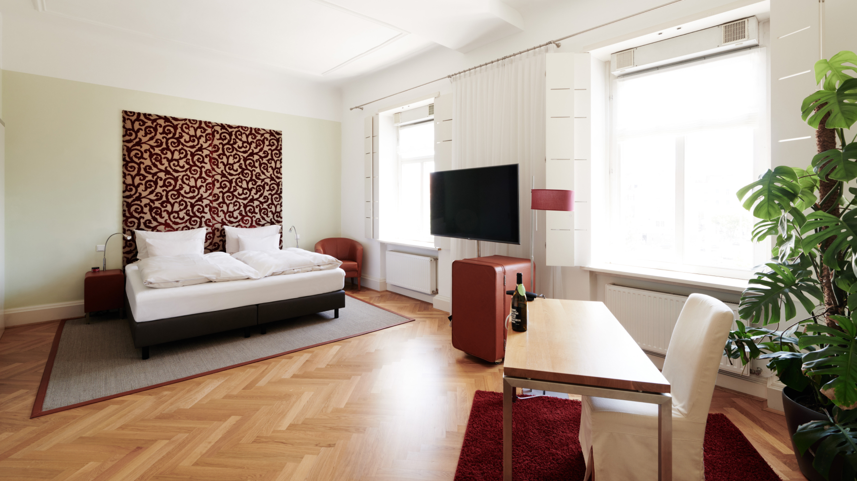 Helles Hotelzimmer mit Bett, Fernseher, Schreibtisch, Fenster und großer Zimmerpflanze.