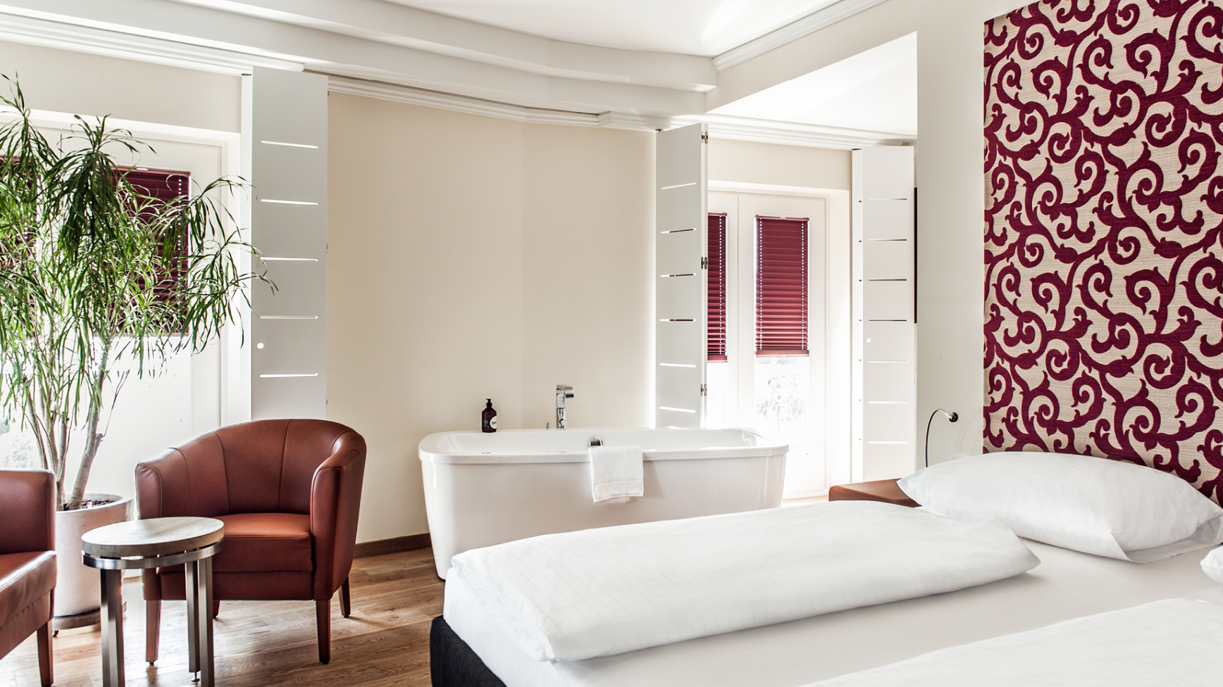 Ein helles Hotelzimmer mit freistehender Badewanne, Sesseln und einem Bett vor einer gemusterten Wand.