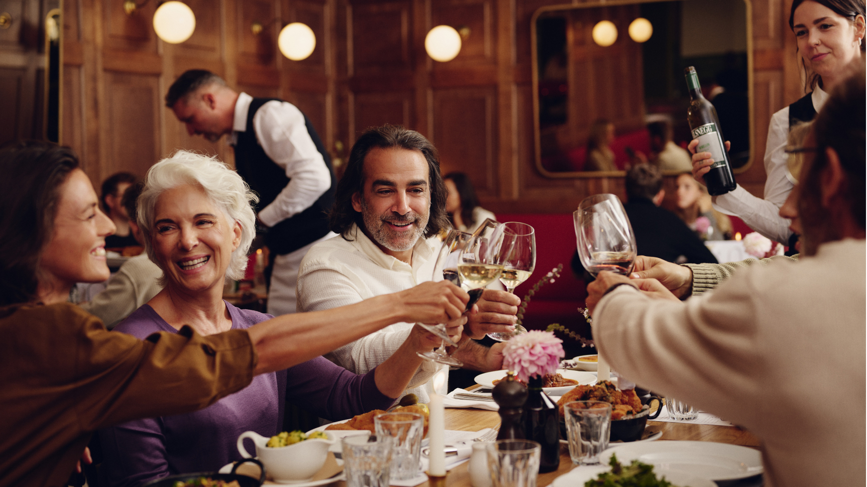 Eine Gruppe von Menschen stößt in einem Restaurant mit Wein an, während ein Kellner bedient.