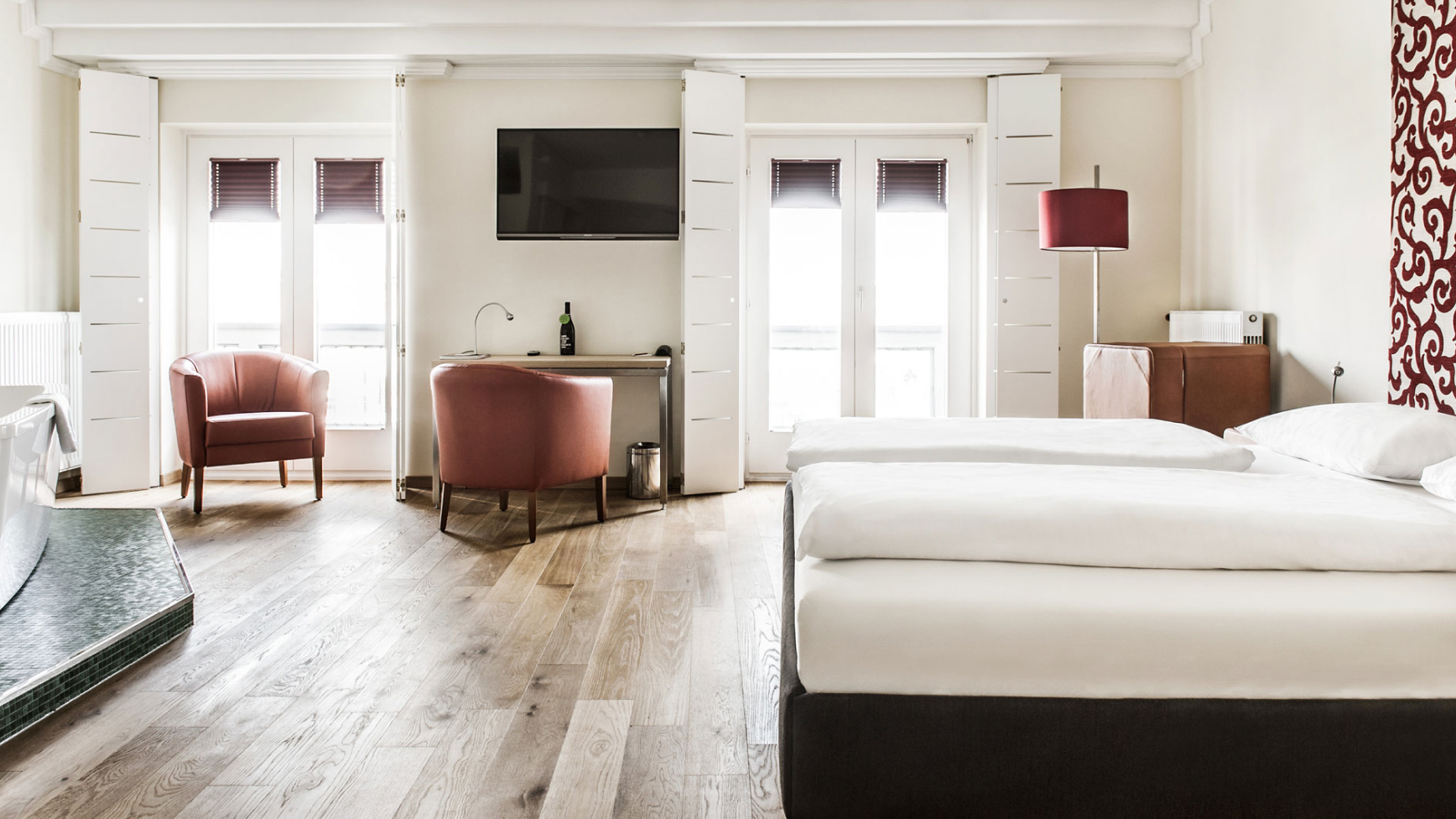 Helles Hotelzimmer mit zwei Sesseln, Schreibtisch, Fernseher, Doppelbett und großen Fenstern.