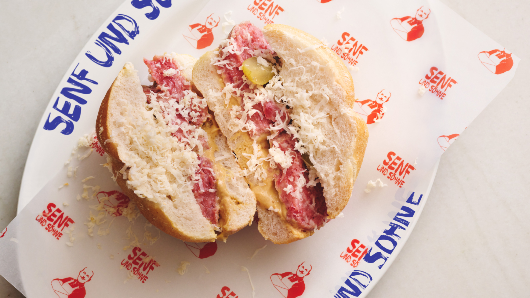 Halbiertes Brötchen von "Senf und Söhne" mit Wurst, Käse und Essiggurken.