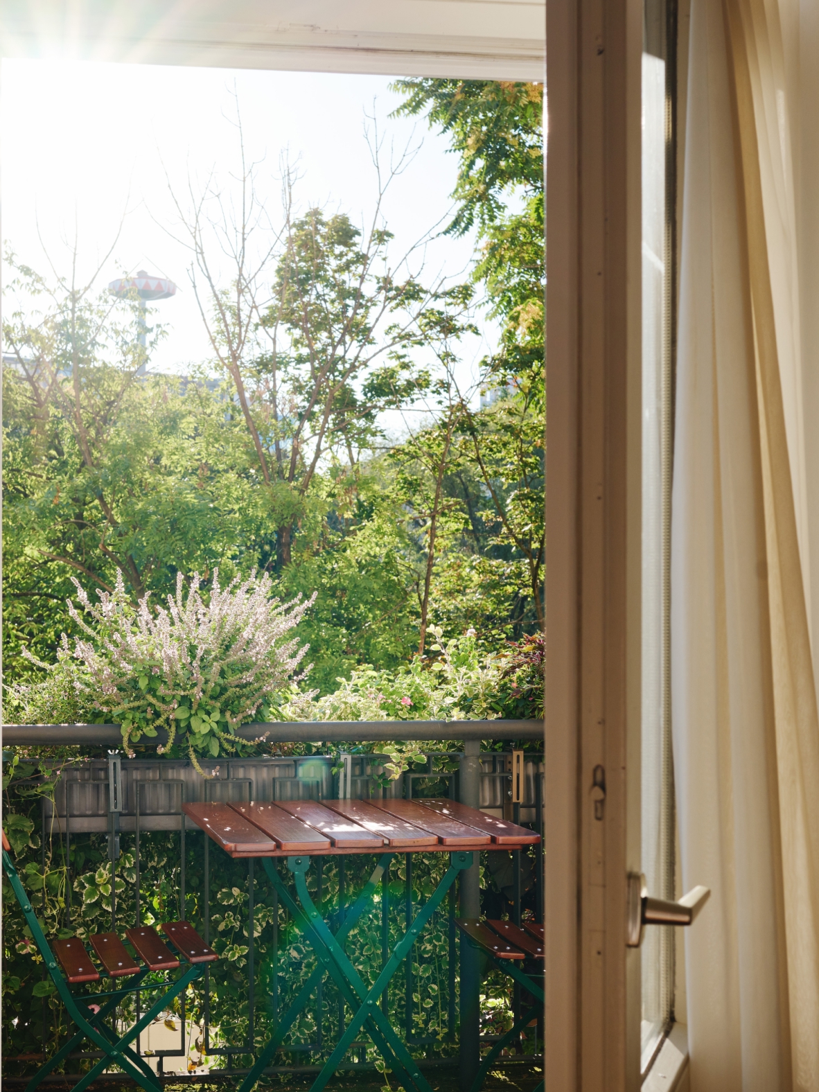 Balkonblick durch geöffnete Tür, Tisch und Stühle. Grünes Laub und im Hintergrund ein Karussell.