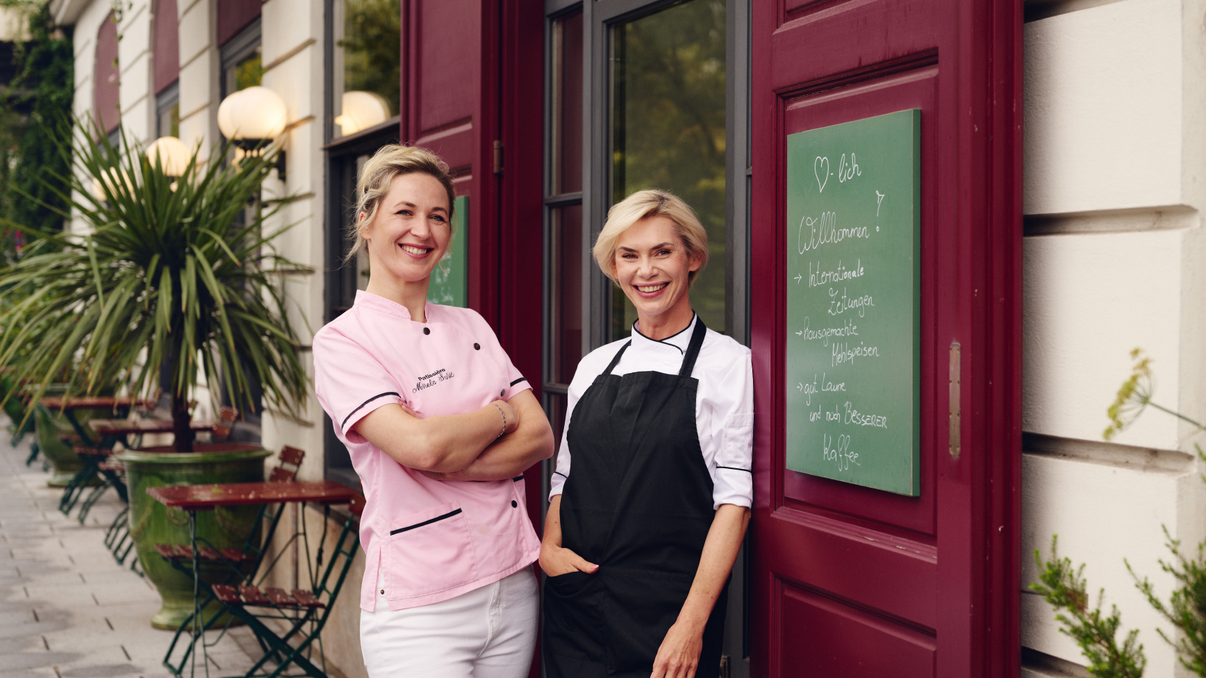 Zwei Frauen stehen vor dem Restaurant „Martha Bäckerei“. Eine Tafel listet Zeitungen, Häppchen, etc. auf.