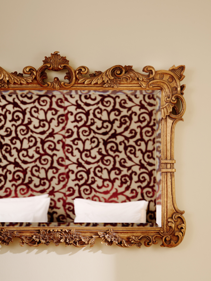 Vergoldeter, kunstvoller Spiegel reflektiert ein rotes, gemustertes Kopfteil und weiße Kissen.