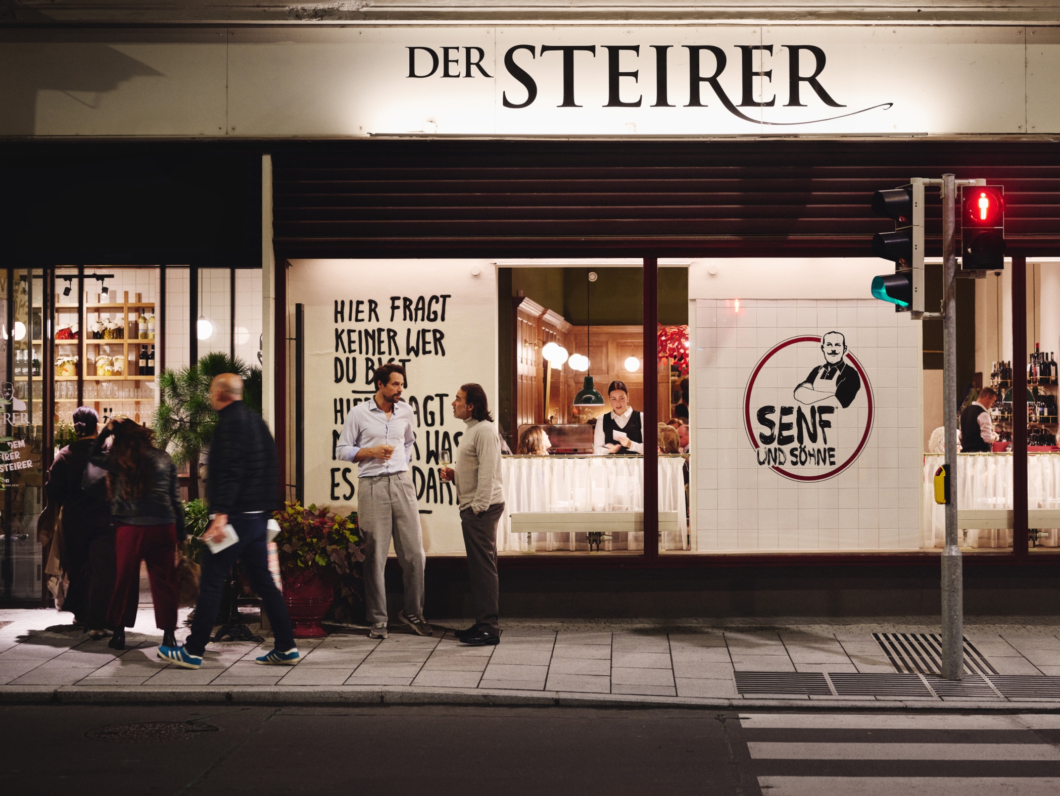 Nachtsaufnahme von "Der Steirer" mit "Senf und Söhne"-Logo; Passanten vor einem beleuchteten Geschäft.