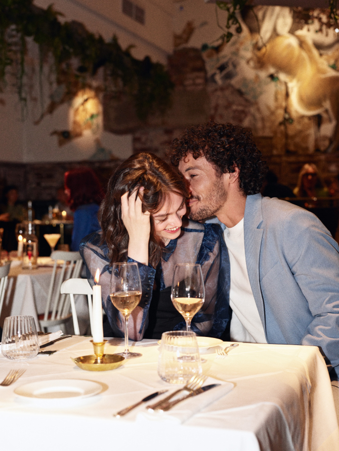 Paar sitzt an gedecktem Tisch in einem Restaurant. Er flüstert ihr ins Ohr, sie lacht.