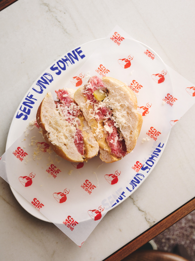 Auf einem Teller von Senf und Söhne liegt ein halbiertes Sandwich mit Fleisch, Käse und Gurken.