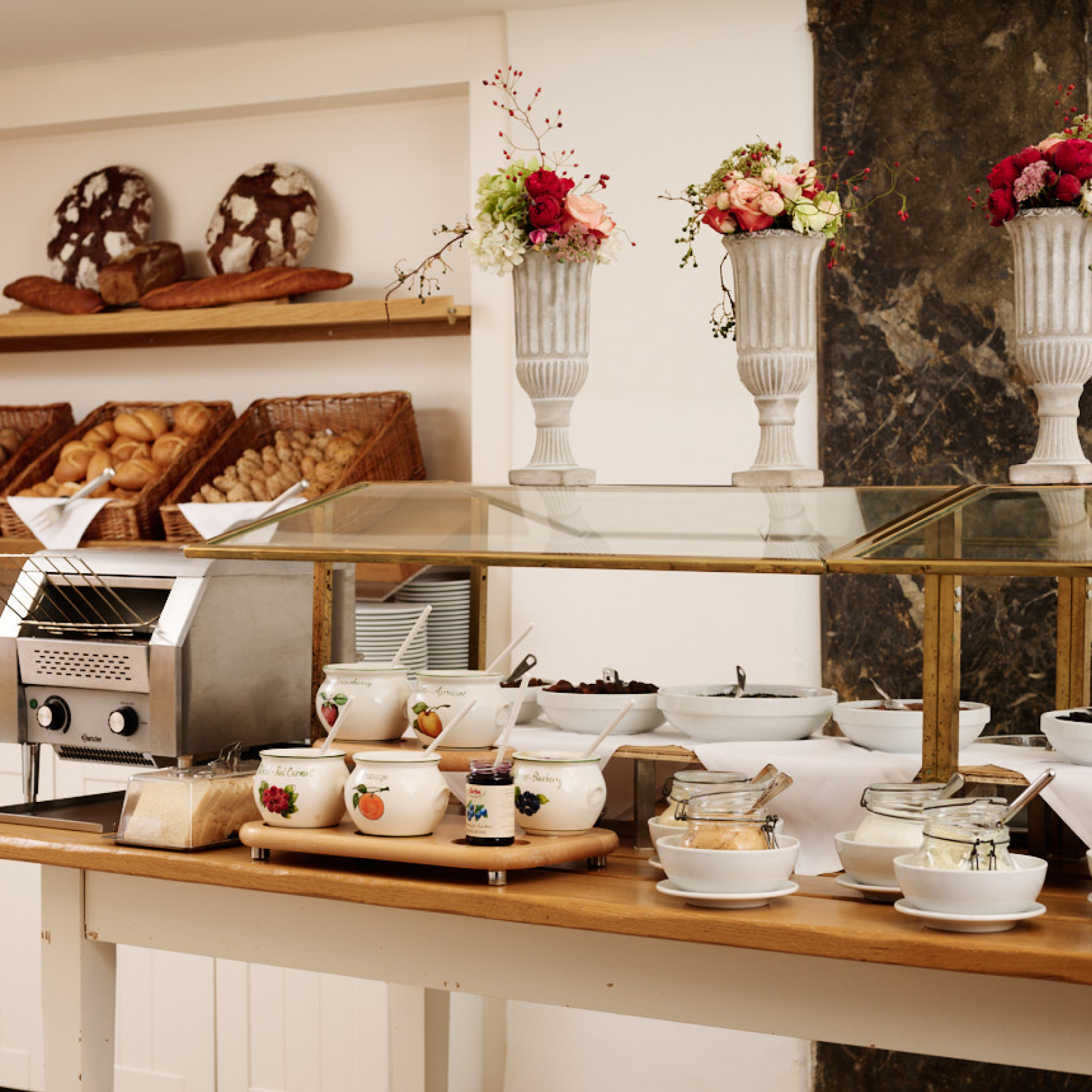 Ein Buffet-Tisch mit Brotkörben, Toaster, Marmelade, Schalen und Vasen mit Blumen.