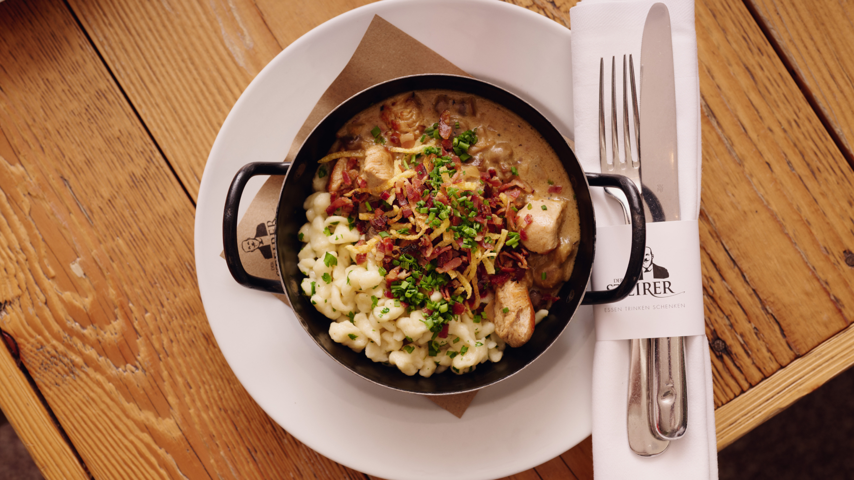 Draufsicht auf Spätzle mit Hühnchen in Pfanne, Besteck in Serviette mit „DER STEIRER“ Logo.