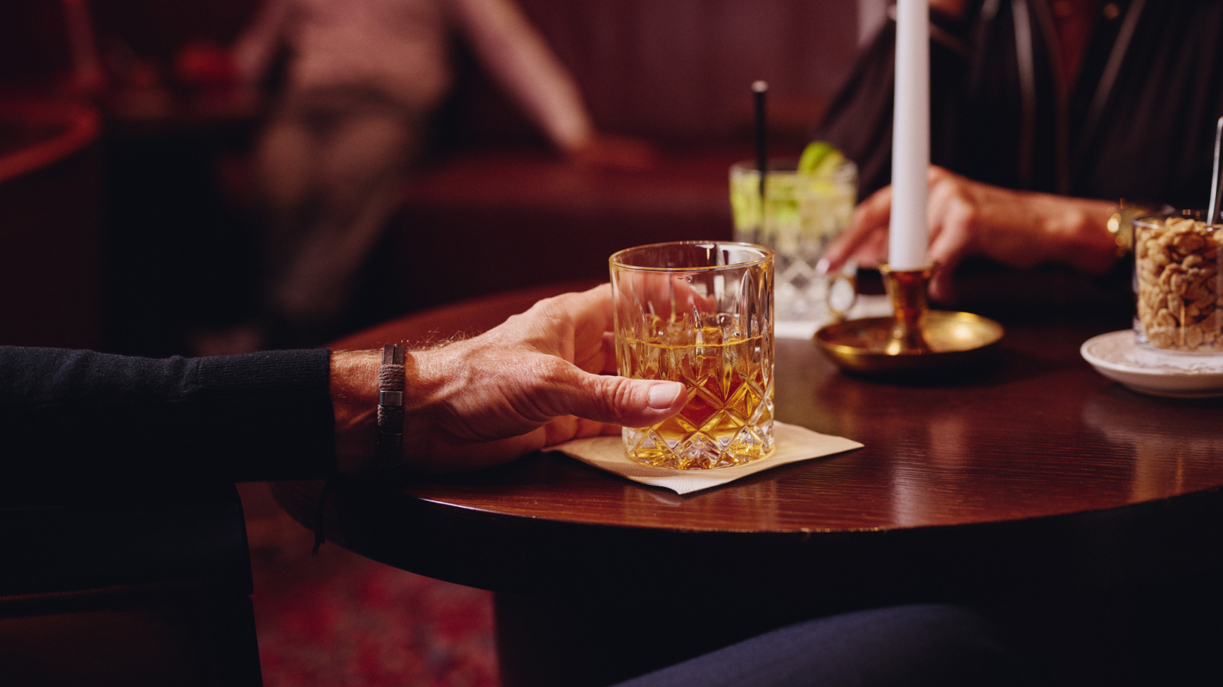 Mann hält Whiskyglas an einem dunklen Tisch, Kerze, Nüsse und anderes Getränk in der Nähe.