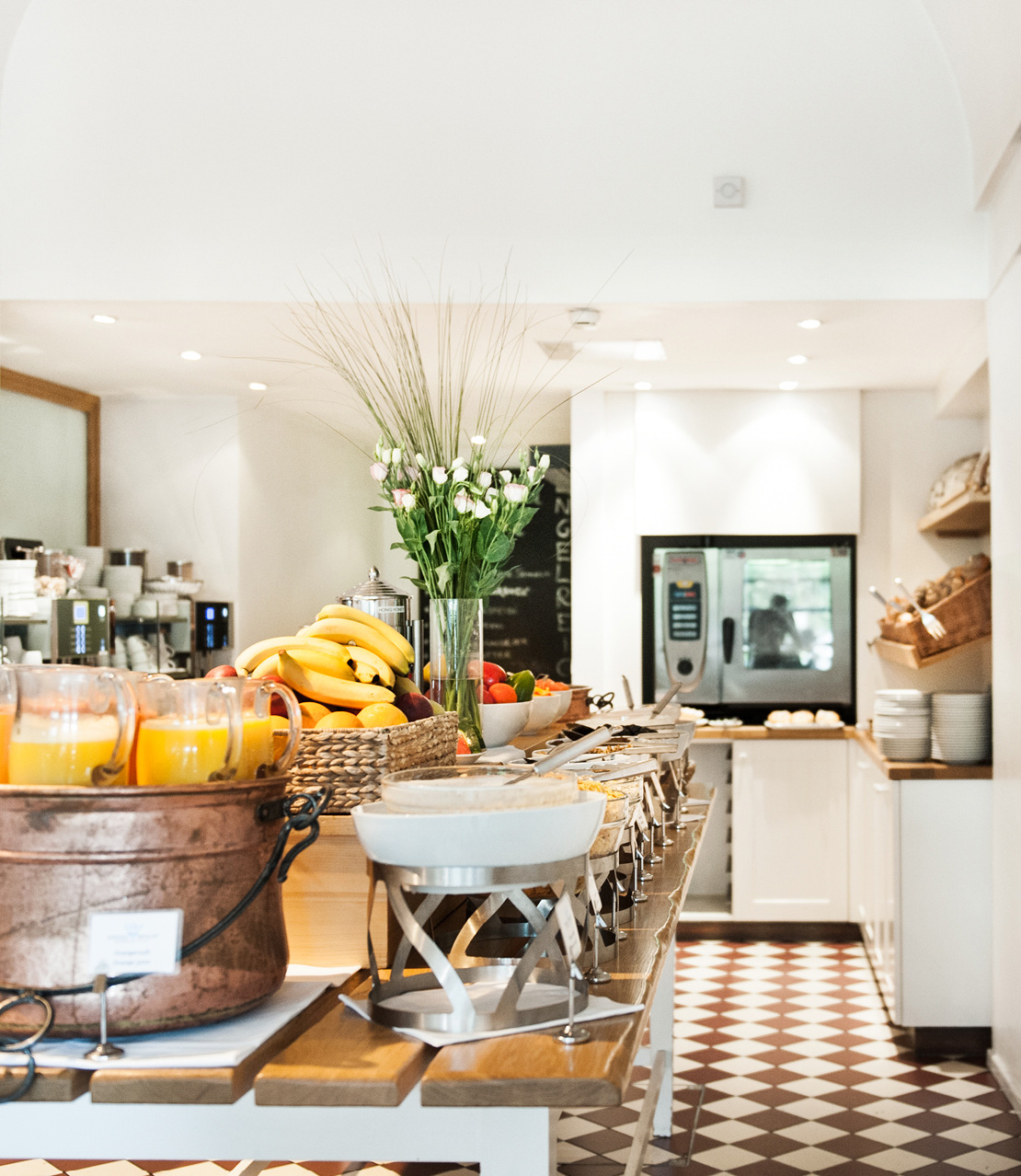 Frühstücksbuffet mit Obst, Bananen, Saftkrügen und Blumen in einem Restaurant. Schachbrettartiger Boden.