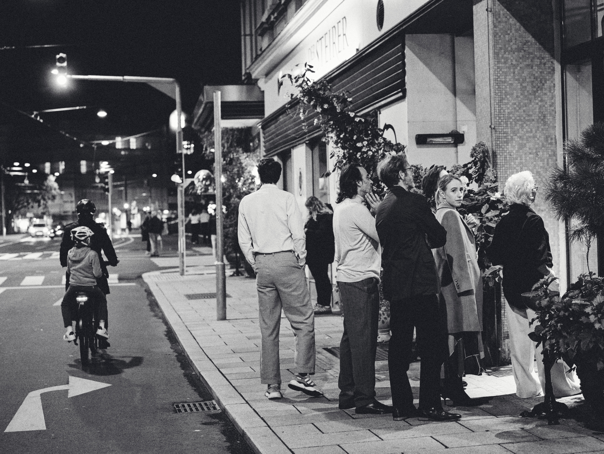 Schwarzweißfoto von Personen vor einem Laden namens "STEINER" bei Nacht. Ein Radfahrer im Vordergrund.