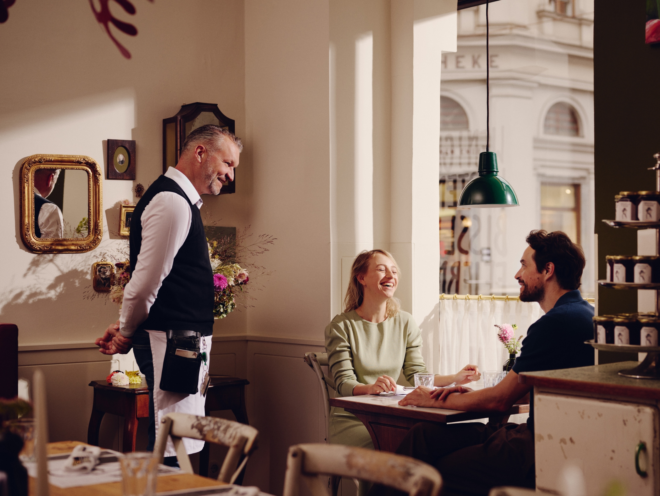 Ein Paar lacht in einem Restaurant, während ein Kellner daneben steht. Im Fenster steht "APOTHEKE".