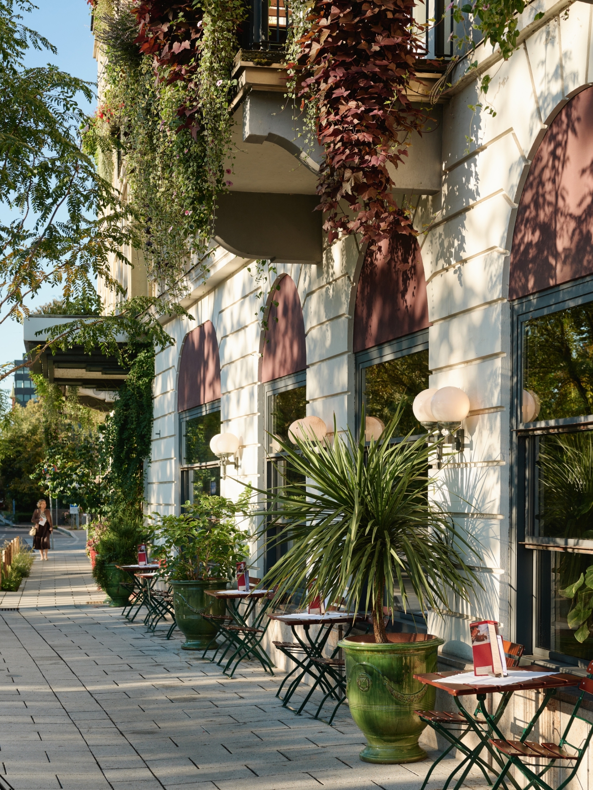 Außenansicht eines Gebäudes mit Pflanzen, Fenstern und Tischen mit Stühlen auf dem Bürgersteig.