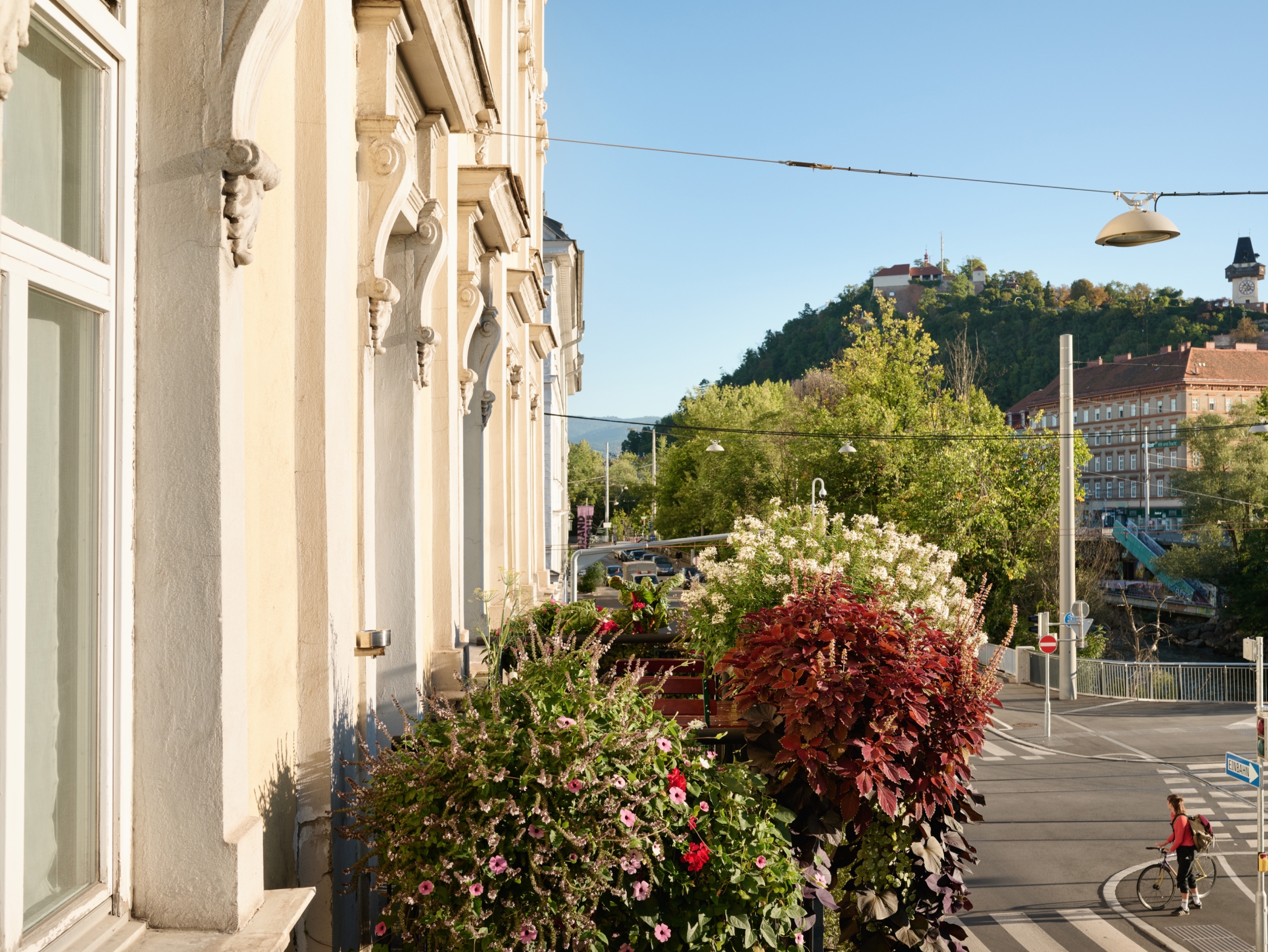 Blick von einem Balkon in Graz auf den Schlossberg mit dem Grazer Uhrturm. Radfahrerin quert Straße.