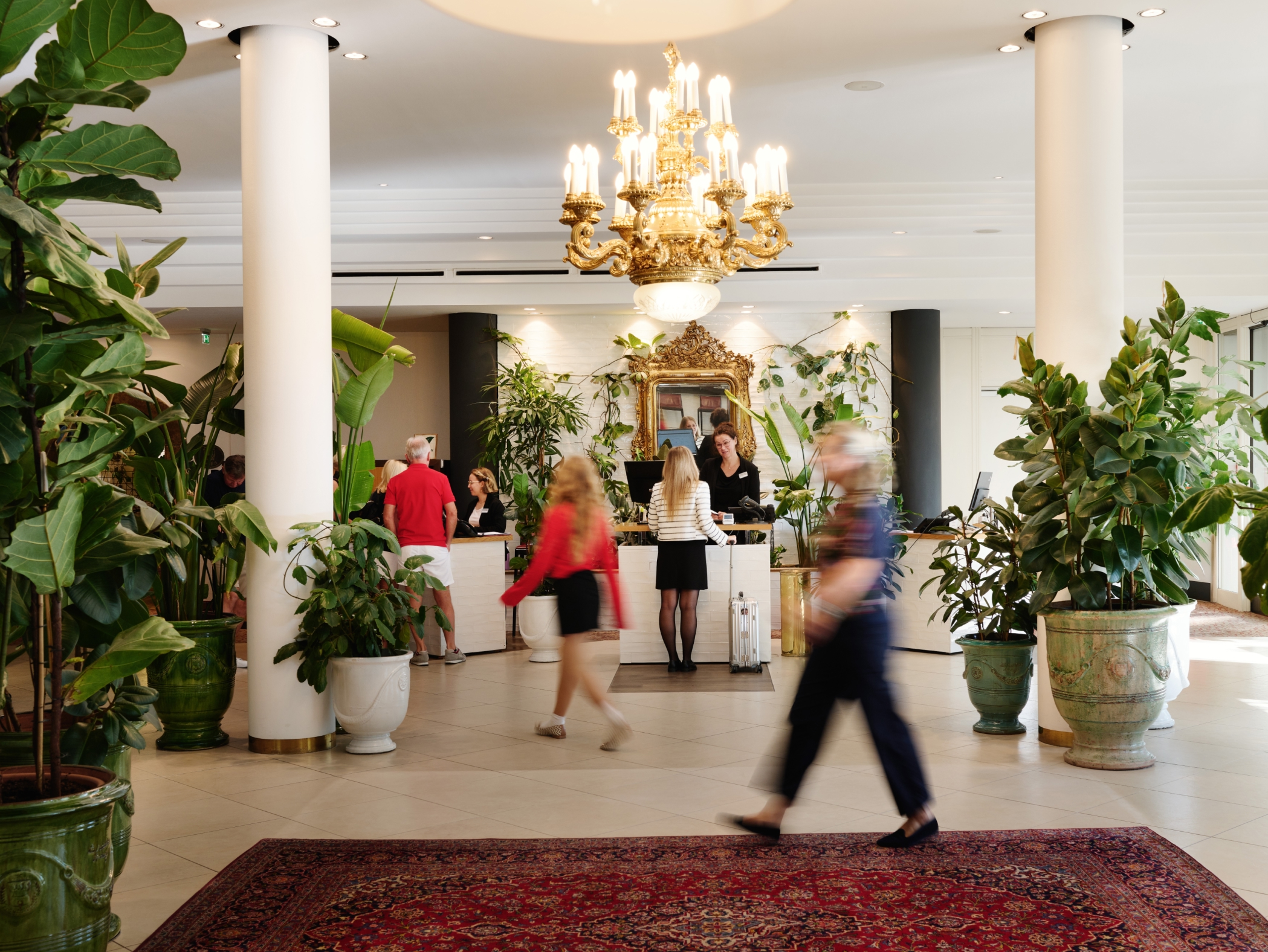 Eingangshalle eines Hotels mit goldenem Kronleuchter, Säulen, Pflanzen und Teppich. Menschen bewegen sich.