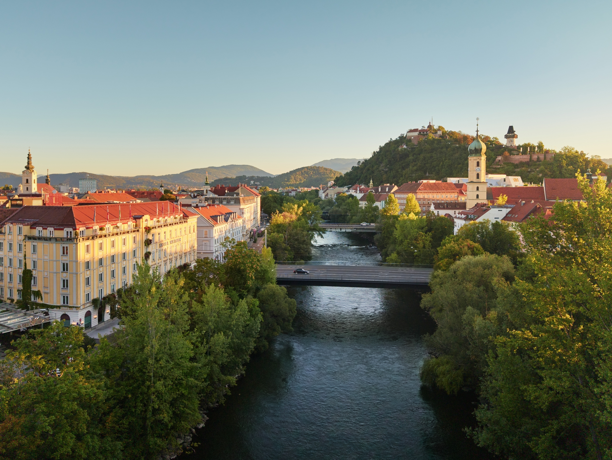 Panorama von Graz, Österreich, mit dem Fluss Mur, Brücken, Gebäuden und einem Hügel mit einem Uhrturm.