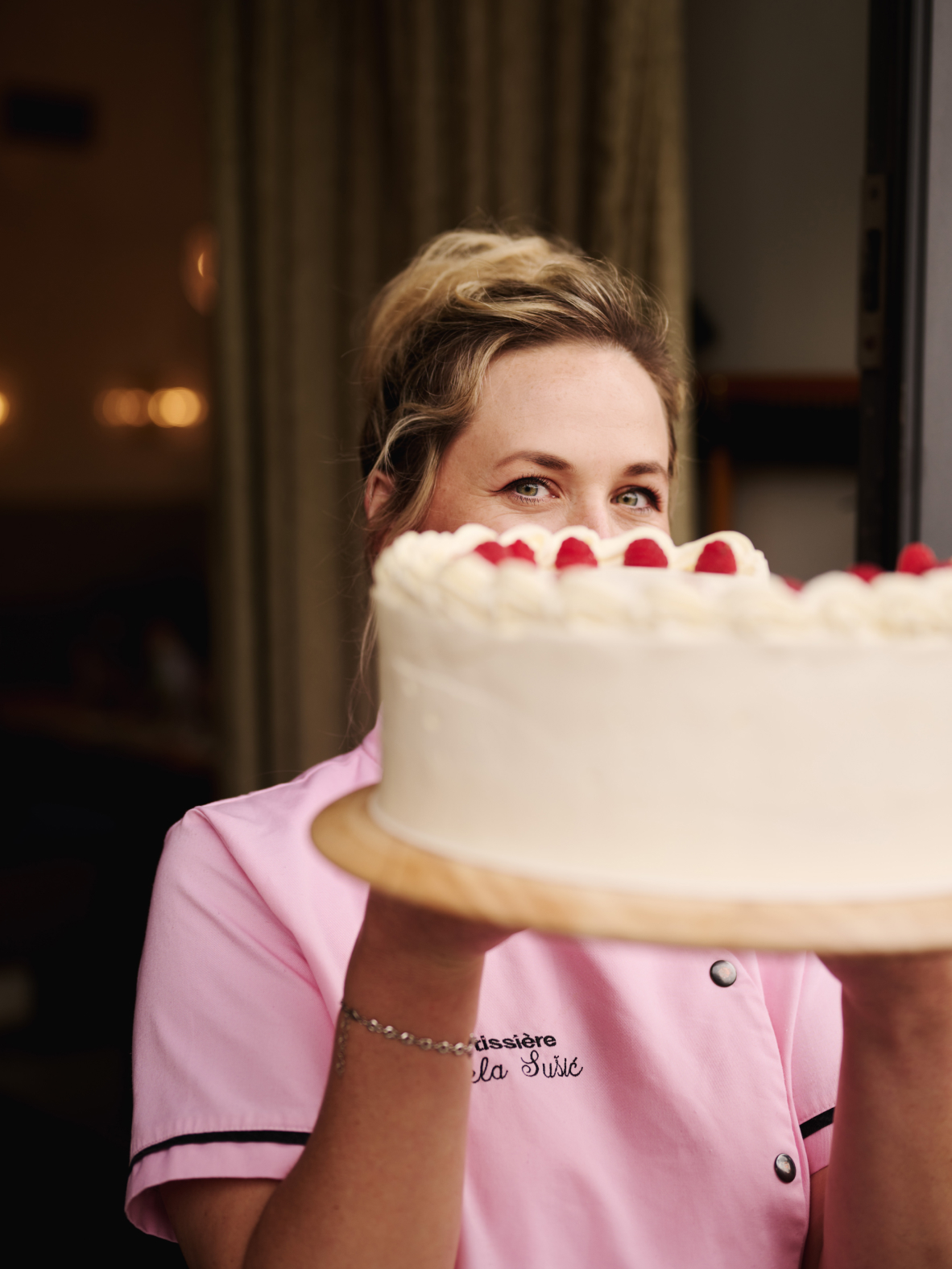 Eine Frau in rosa Uniform hält eine Sahnetorte mit Himbeeren hoch. "Pâtissière à la Susie" steht auf der Uniform.