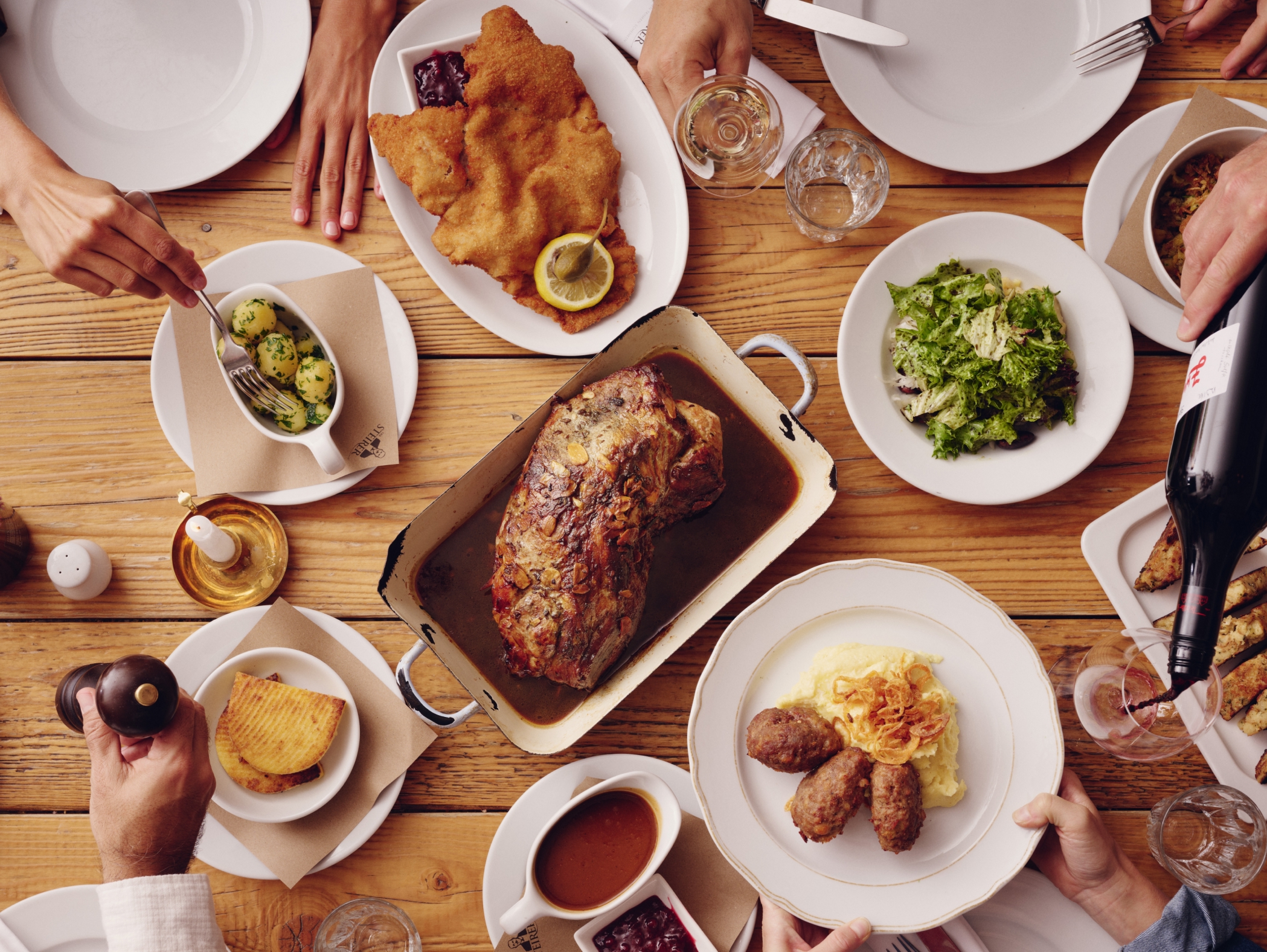 Draufsicht: Holztisch mit Wiener Schnitzel, Braten, Salat, Kartoffeln und Person, die Wein einschenkt.