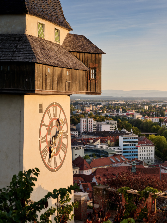 Der Grazer Uhrturm mit Stadtblick, der Uhrturm hat eine große Uhr mit römischen Ziffern.