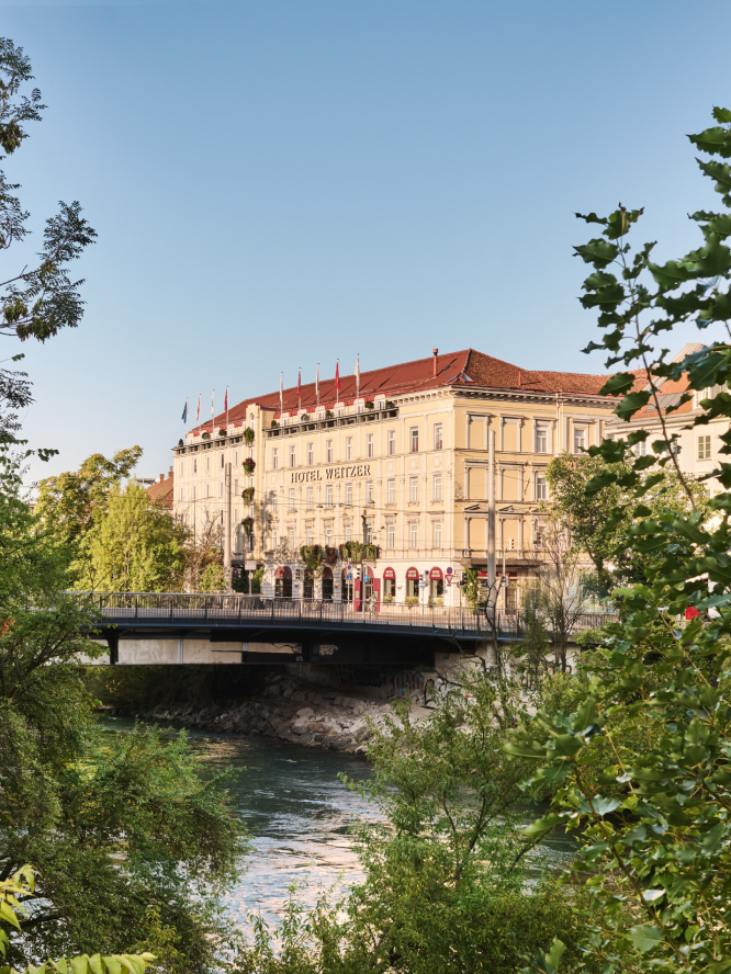 Ansicht des Hotel Weitzer in Graz über einen Fluss mit Brücke. Umgeben von viel Grün.