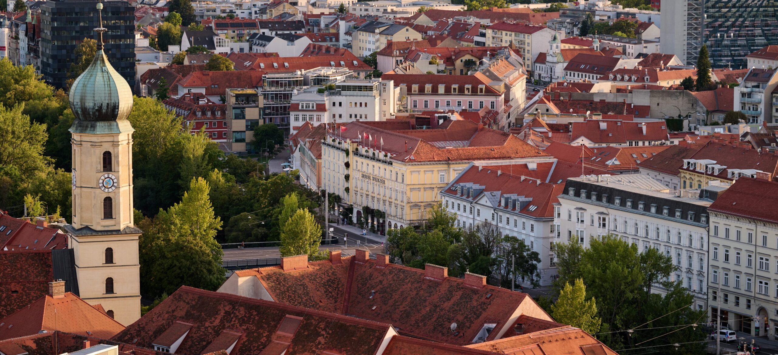 Luftaufnahme von Graz, Österreich, mit Blick auf Gebäude mit roten Ziegeldächern und Kirchturm.